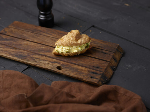 Chopped egg salad croissant sandwich with lettuce on a wooden board with brown cloth napkin