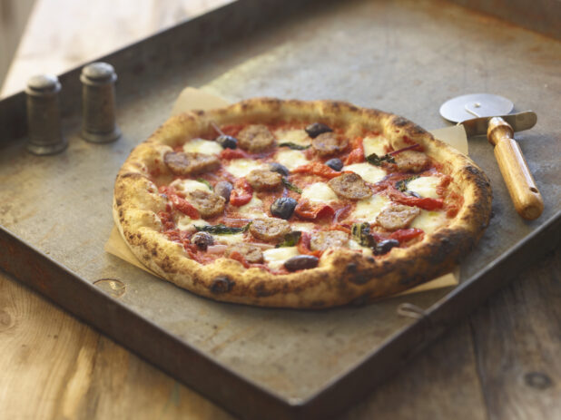 Whole four-topping pizza on a metal board with a pizza cutter and salt and pepper shakers