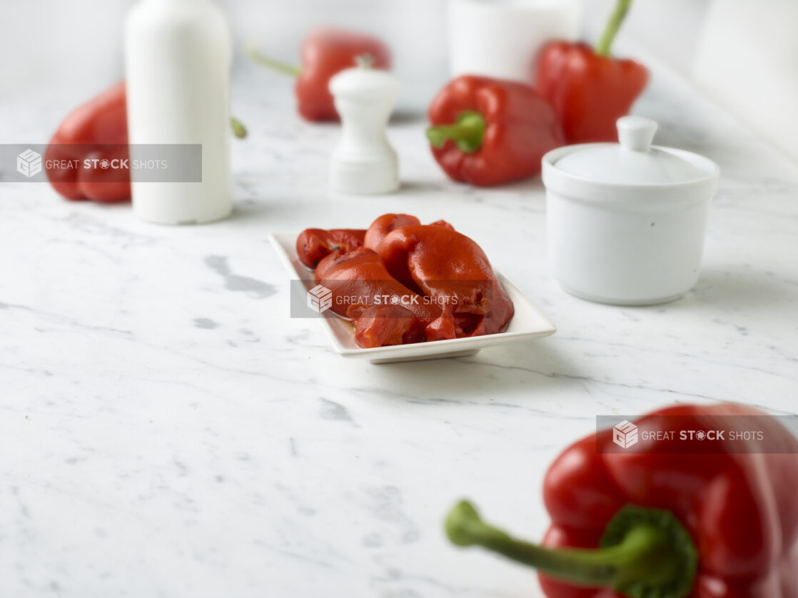 Roasted red peppers on a small rectangular plate with fresh red bell peppers around it on a white marble background