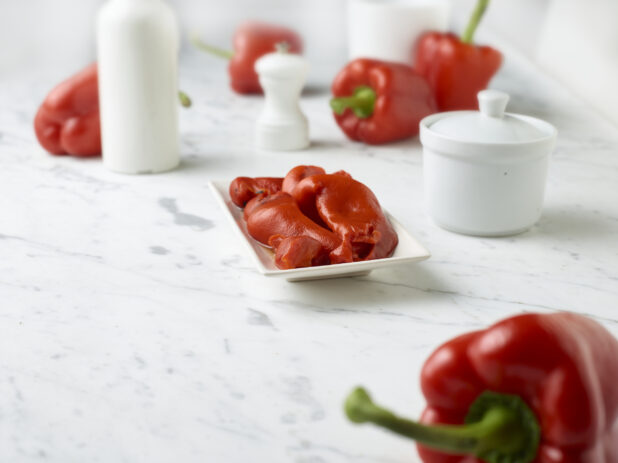Roasted red peppers on a small rectangular plate with fresh red bell peppers around it on a white marble background