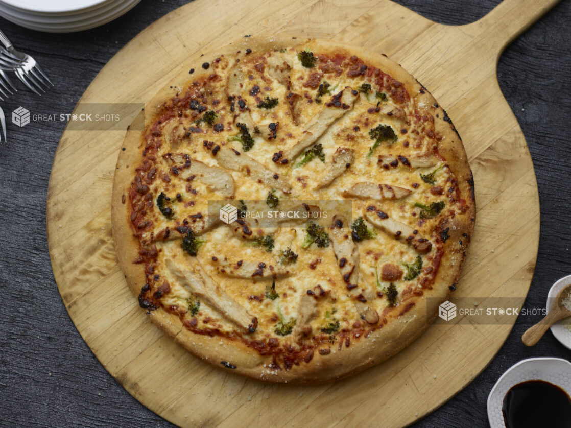 Overhead whole chicken, broccoli, and cheddar pizza on a wooden peel with a black wood background