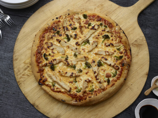 Overhead whole chicken, broccoli, and cheddar pizza on a wooden peel with a black wood background