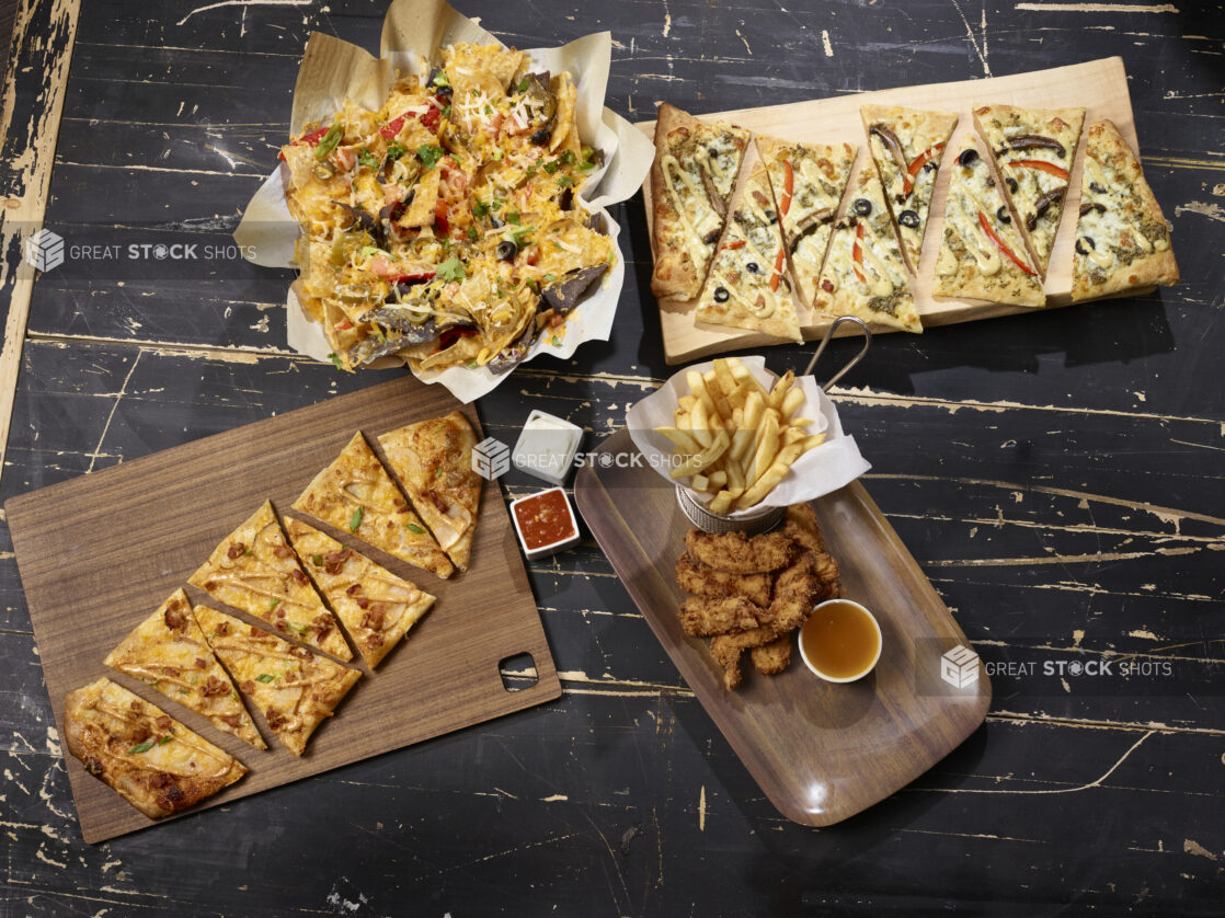 Various appetizers including nachos, flatbreads, chicken tenders and fries, overhead on a dark distressed background