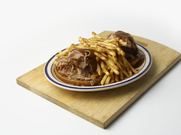 Hot open-faced roast beef sandwich on rye with gravy and french fries on a wooden board on a white background