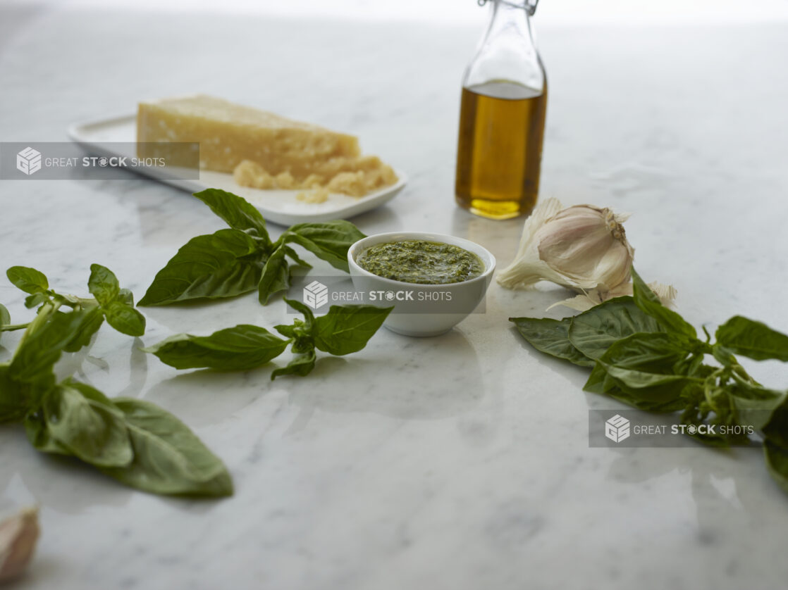 Small white bowl of pesto surrounded by ingredients on white marble