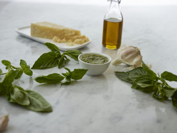 Small white bowl of pesto surrounded by ingredients on white marble