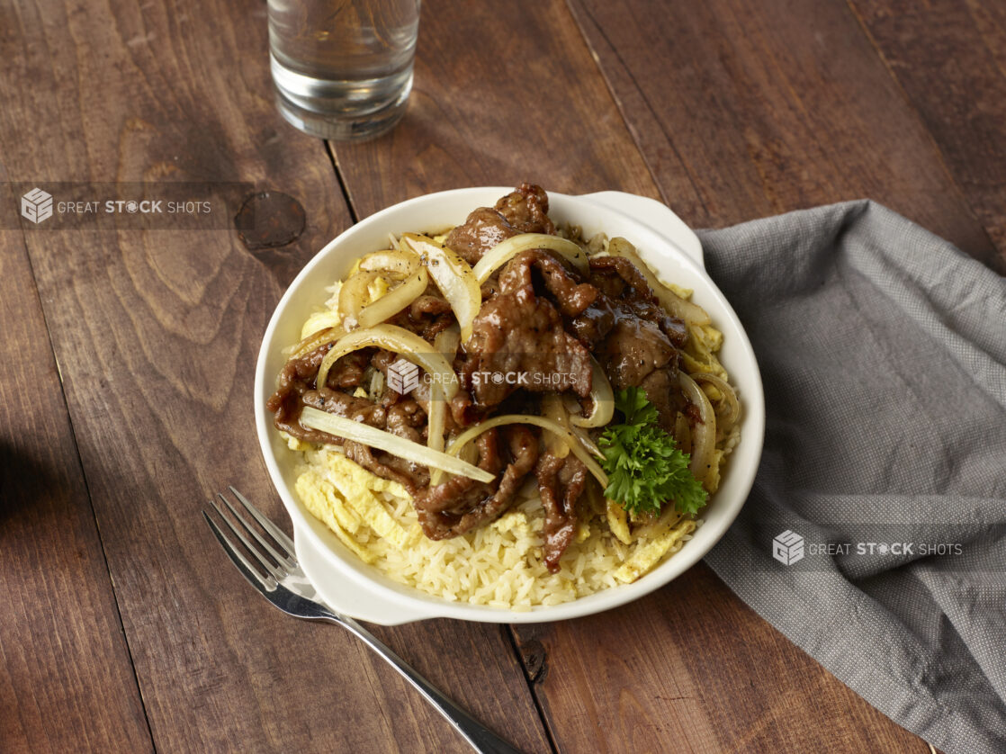 Steak and eggs with sautéed onions on a bed of rice in a white bowl on a wooden background, overhead view