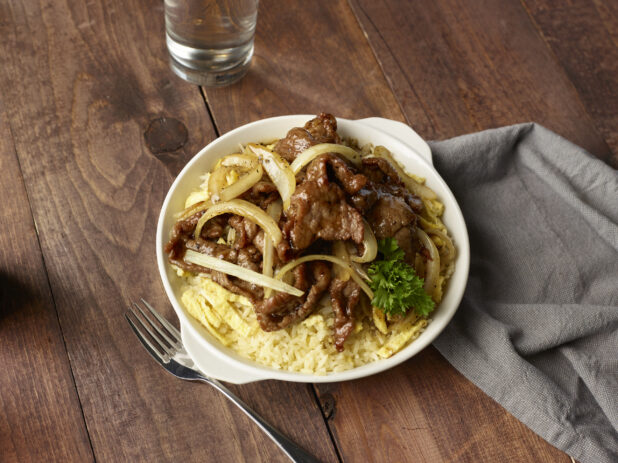 Steak and eggs with sautéed onions on a bed of rice in a white bowl on a wooden background, overhead view