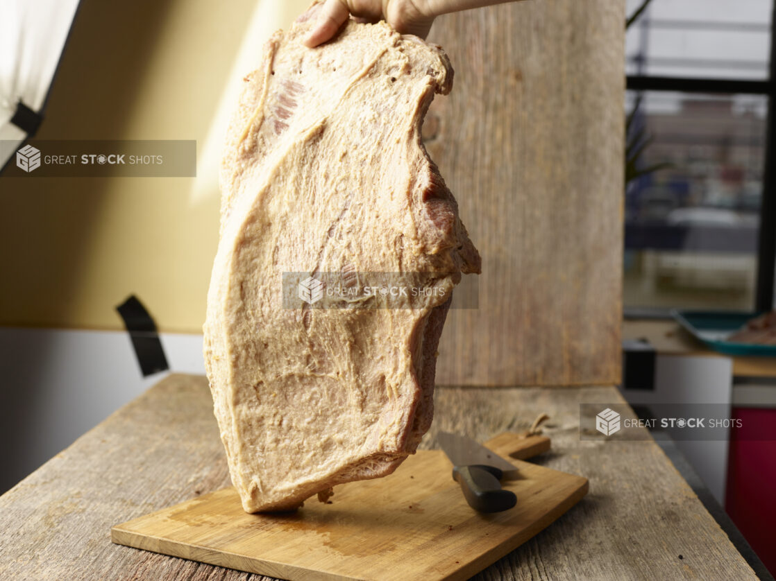 Hand holding a whole guanciale upright on a wood cutting board with a knife on it, commercial setting