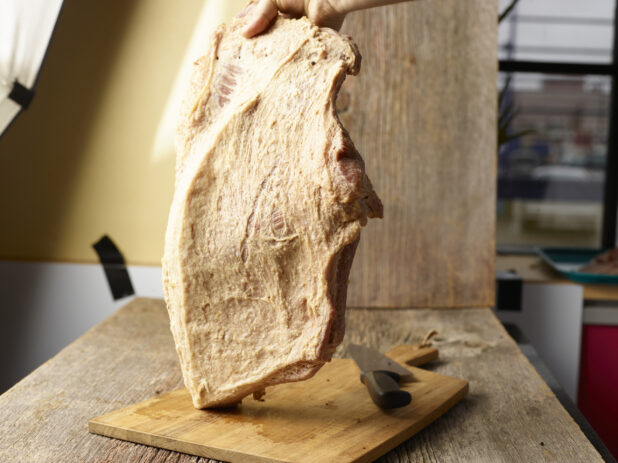 Hand holding a whole guanciale upright on a wood cutting board with a knife on it, commercial setting