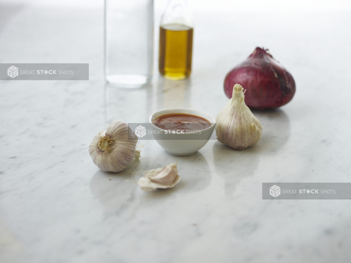 Small white bowl of BBQ sauce surrounded by ingredients on white marble