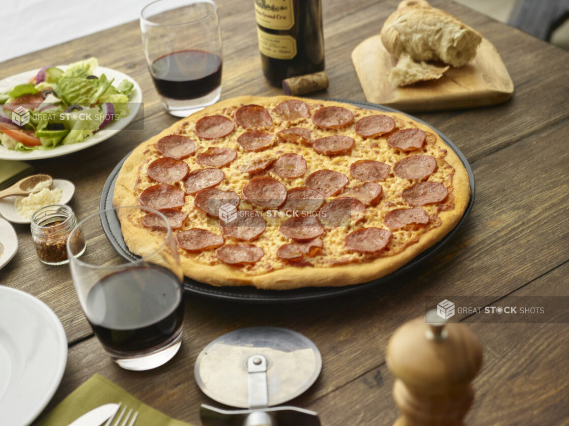 Whole pepperoni pizza on a metal baking pan with red wine and a side salad