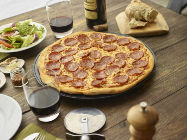 Whole pepperoni pizza on a metal baking pan with red wine and a side salad