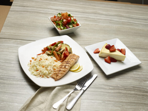 Grilled salmon, rice and sautéed vegetable dish with a side salad and cheesecake for dessert on a wooden background
