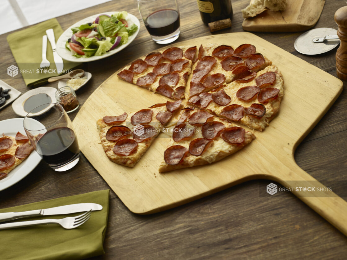 Sliced pepperoni pizza sitting on a peel with a side salad and glasses of red wine