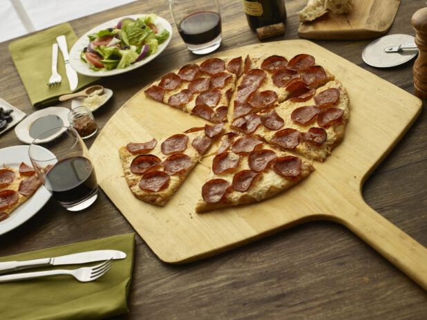 Sliced pepperoni pizza sitting on a peel with a side salad and glasses of red wine