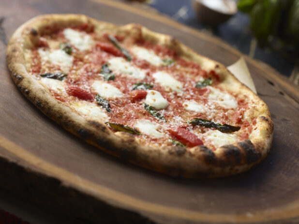 Whole margherita pizza with basil topped with fresh parmesan cheese on a dark wooden board, close-up