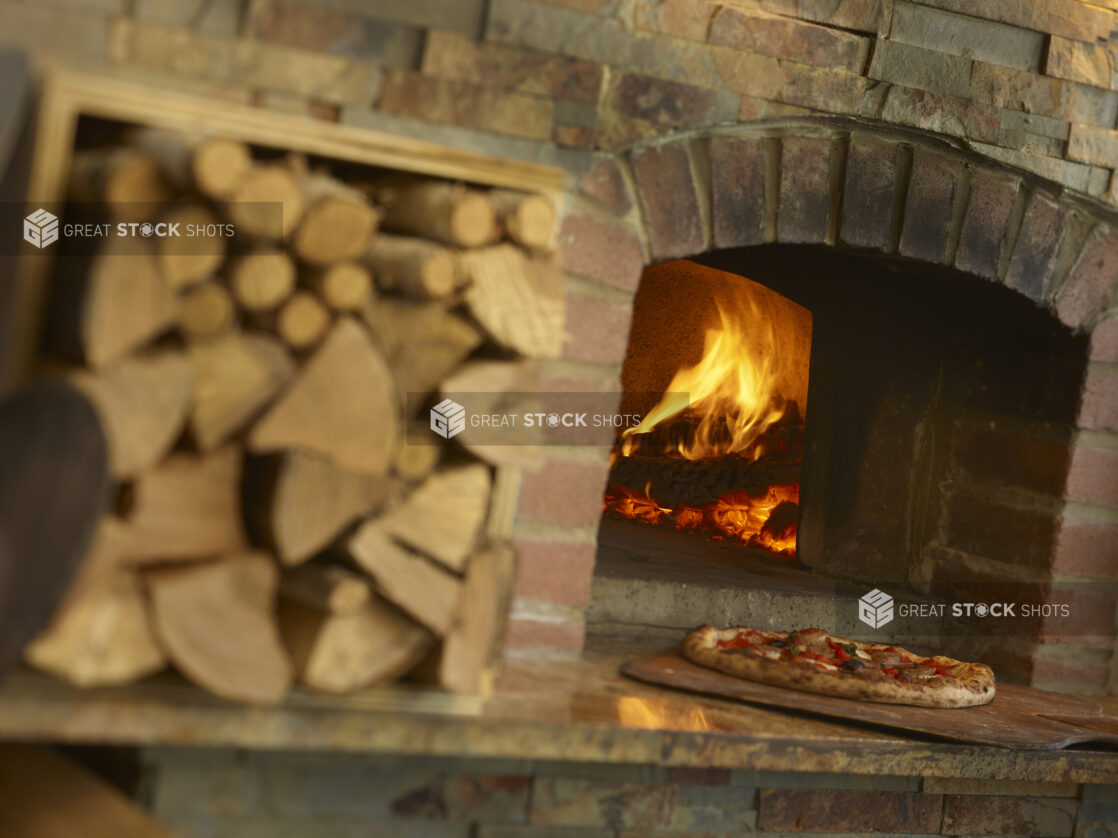 Pizza being taken out of a wood fire oven on a paddle with chopped wood out of focus