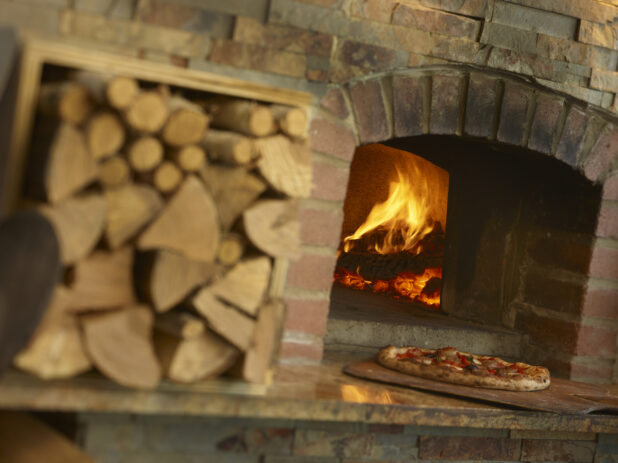 Pizza being taken out of a wood fire oven on a paddle with chopped wood out of focus
