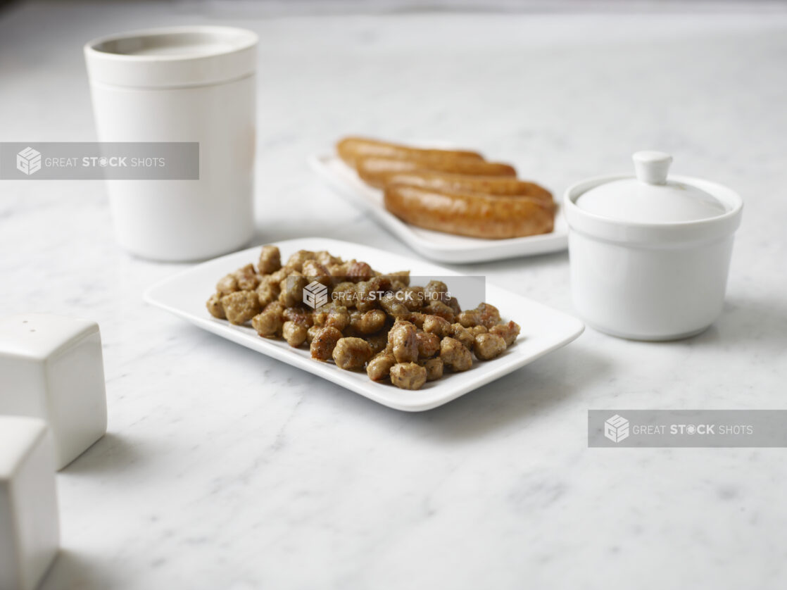 White plate of sausage chunks and full sausage surrounded by white containers on a white marble background