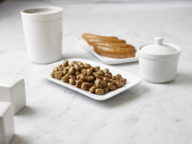 White plate of sausage chunks and full sausage surrounded by white containers on a white marble background