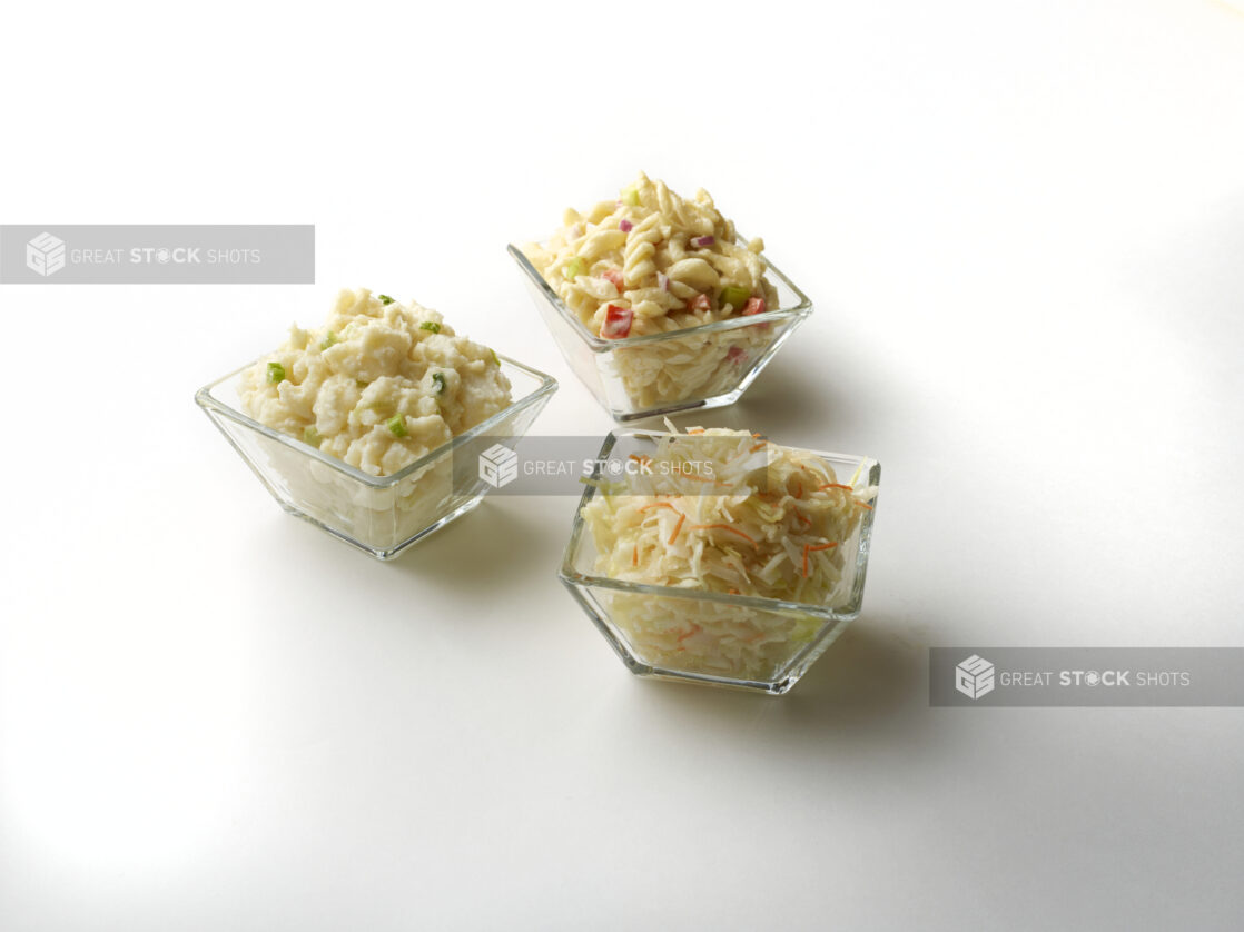 Three glass dishes with cold pasta salad, potato salad and coleslaw on a white background