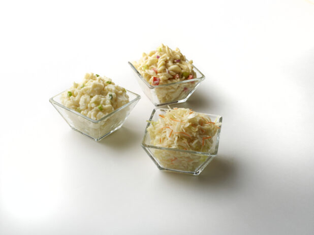 Three glass dishes with cold pasta salad, potato salad and coleslaw on a white background