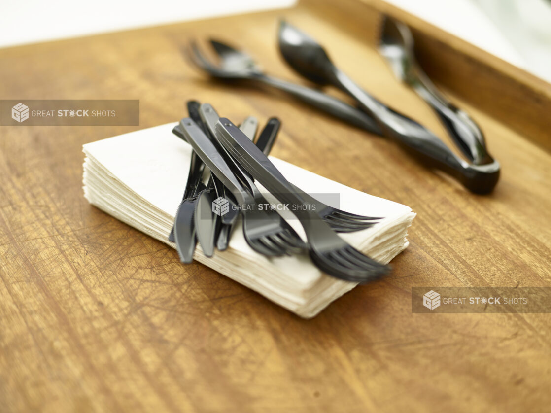 Black disposable plastic cutlery and utensils with paper napkins on a wooden cutting board