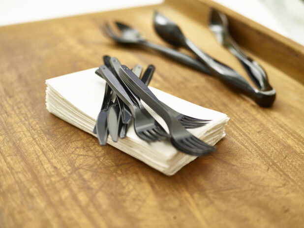 Black disposable plastic cutlery and utensils with paper napkins on a wooden cutting board