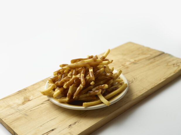 Plate of shoestring french fries covered in gravy on a wooden board with a white background