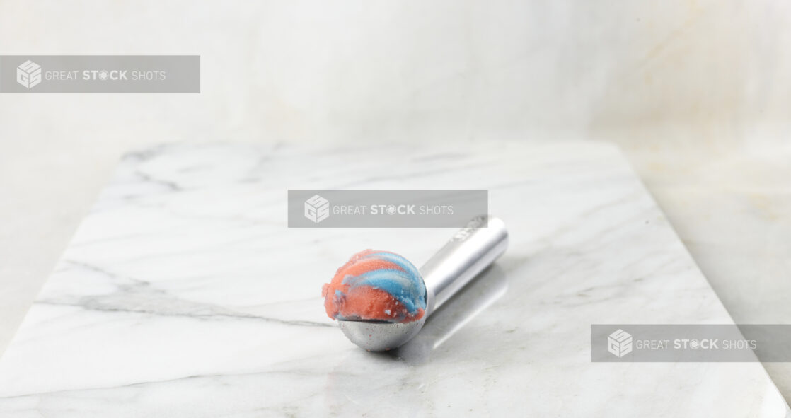 Scoop of rainbow gelato on a metal scoop with a white marble background