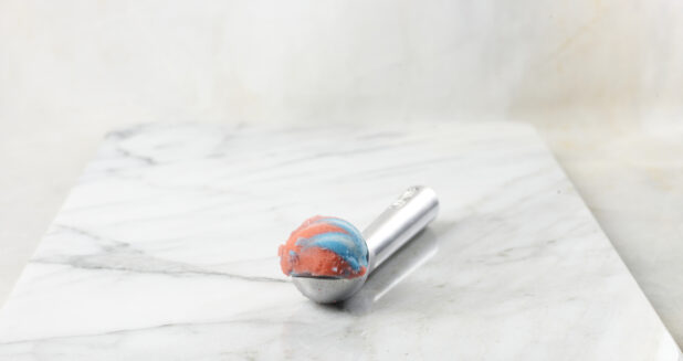 Scoop of rainbow gelato on a metal scoop with a white marble background