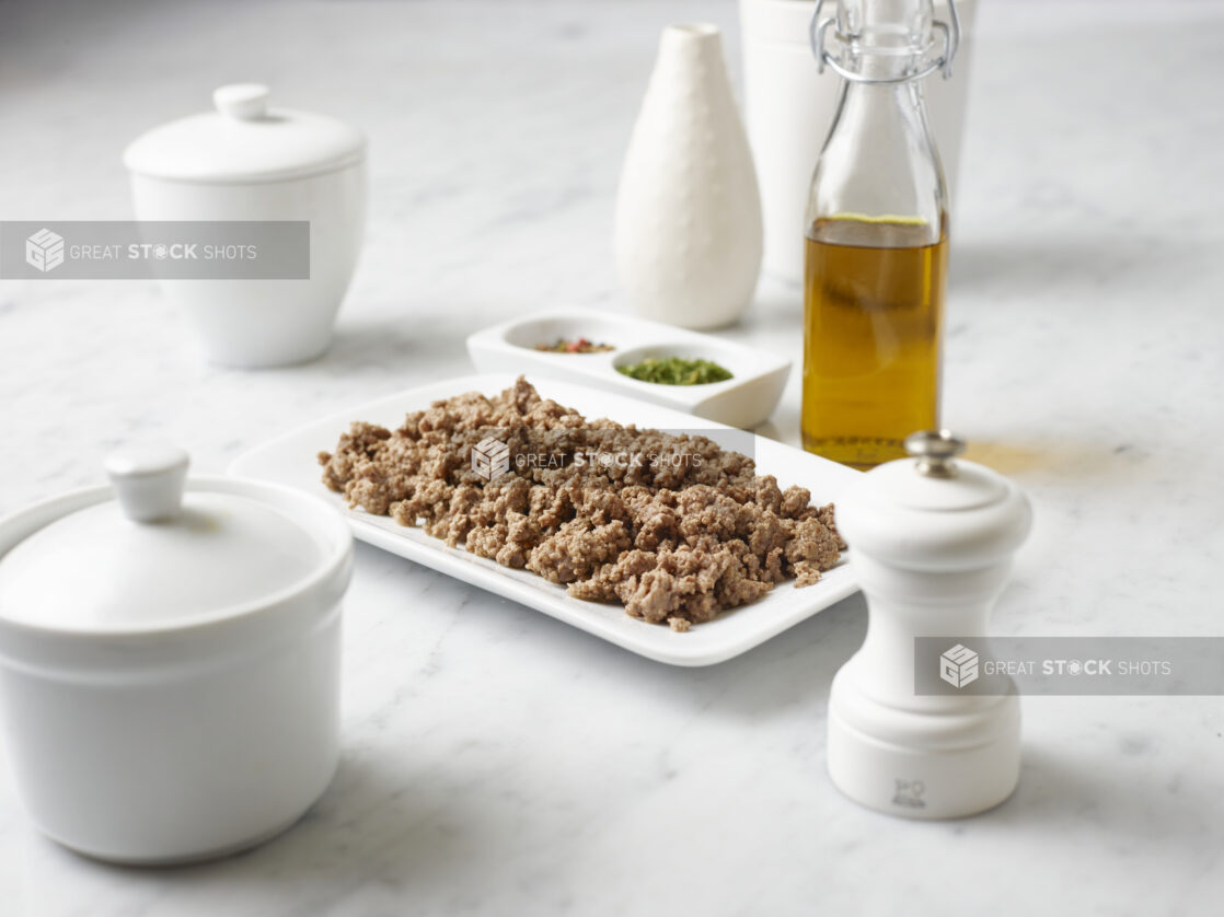 White plate of ground beef surrounded by ingredients and white jars on a white marble background