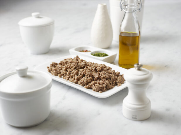 White plate of ground beef surrounded by ingredients and white jars on a white marble background