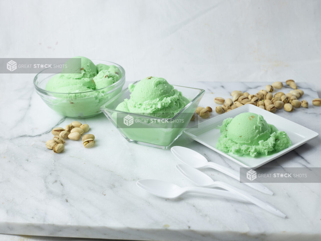 Three bowls of pistachio ice cream surrounded by unshelled pistachio nuts and spoons on a white marble background