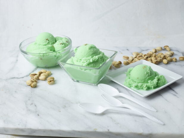 Three bowls of pistachio ice cream surrounded by unshelled pistachio nuts and spoons on a white marble background