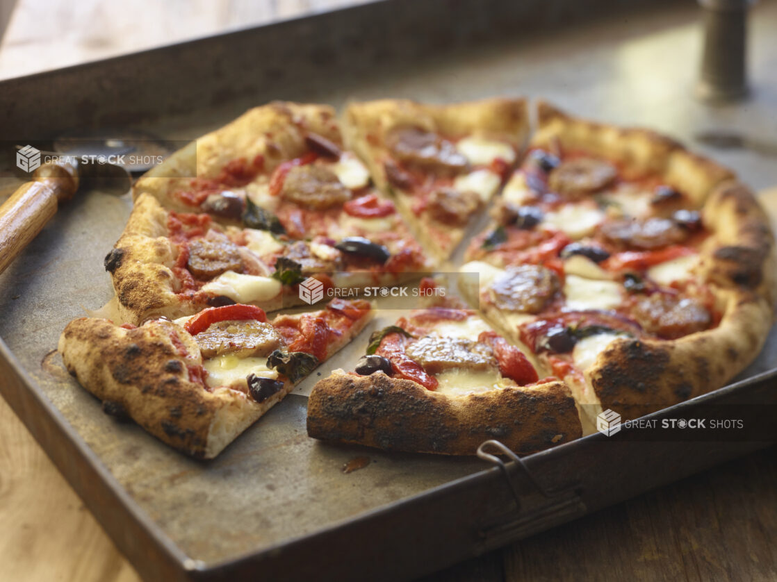 Sliced three-topping pizza with sausage, roasted red pepper, and black olives on a metal board with a pizza cutter