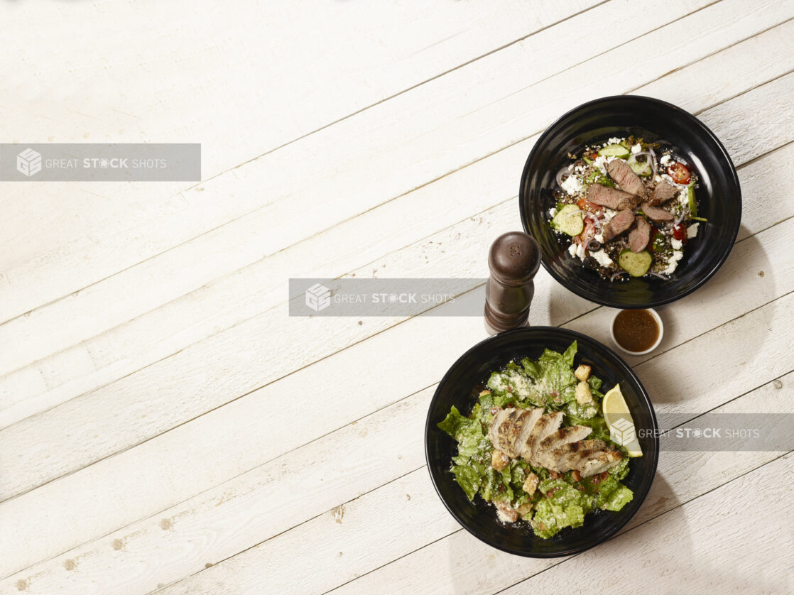 Overhead of caesar salad with chicken and greek salad with steak on a light wood background