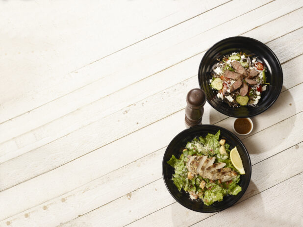 Overhead of caesar salad with chicken and greek salad with steak on a light wood background