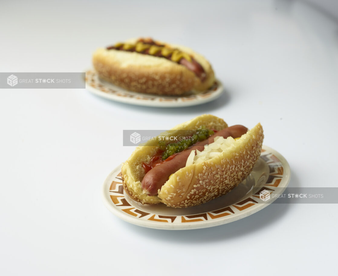 Two loaded hot dogs, one with relish, ketchup, mustard and onions on a sesame seed bun on a white background, close-up