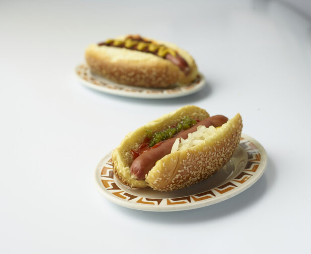 Two loaded hot dogs, one with relish, ketchup, mustard and onions on a sesame seed bun on a white background, close-up