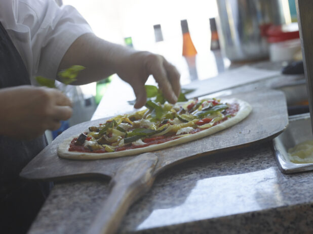 Pizza chef sprinkling fresh basil on a raw pizza with grilled vegetables on a peel in a restaurant