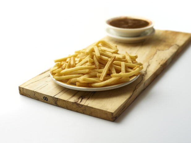 Plate of plain french fries with a bowl of soup in the background on a wooden board with a white background