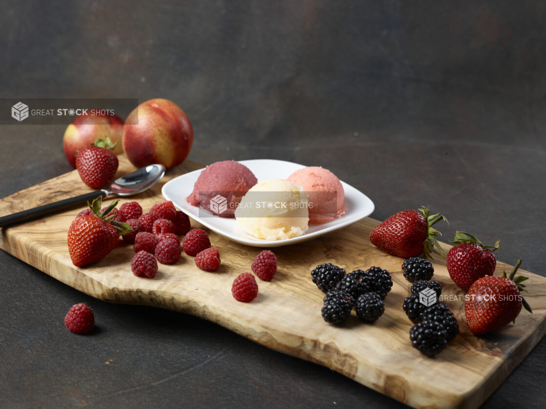 Three scoops of gelato on a white plate surrounded by fresh fruit on a wooden board with a dark background