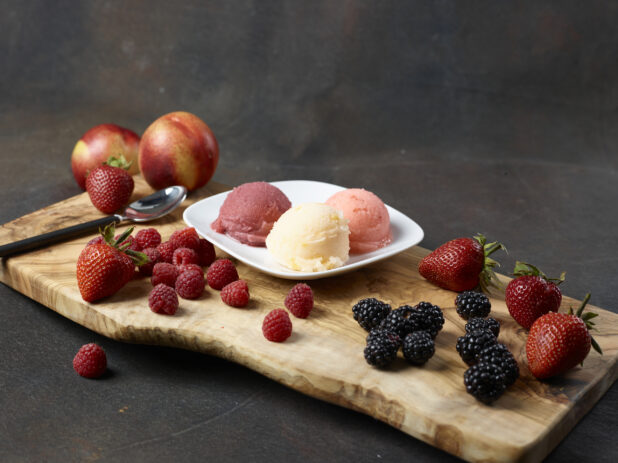 Three scoops of gelato on a white plate surrounded by fresh fruit on a wooden board with a dark background