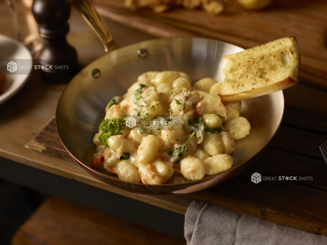 Gnocchi in a creamy spinach and roasted red pepper sauce with garlic bread on the side in a steel saucepan on a wooden table