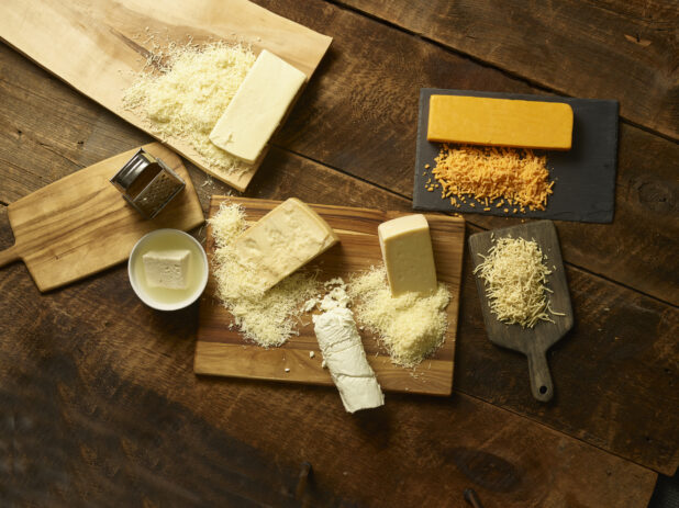 Overhead of various types of cheese, shredded / grated on wooden boards on a dark wooden background