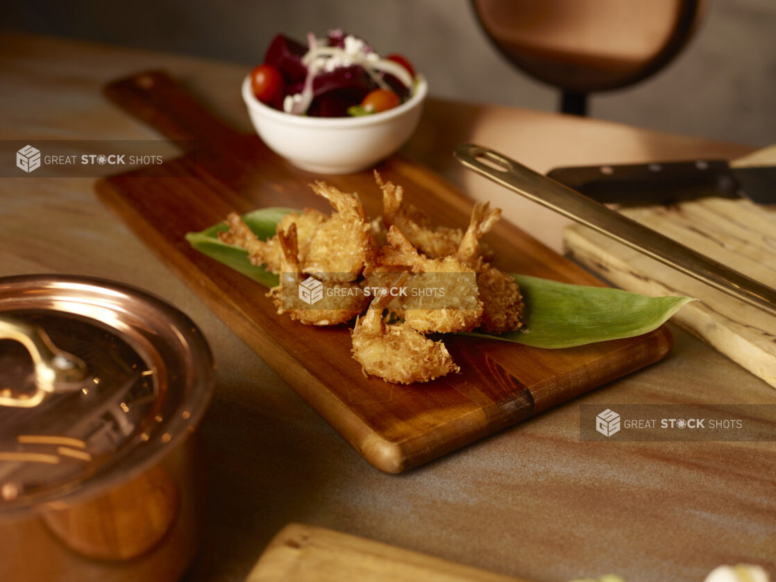 Coconut shrimp piled on a long green leaf on a wooden board with a side beet salad