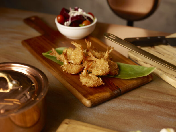 Coconut shrimp piled on a long green leaf on a wooden board with a side beet salad