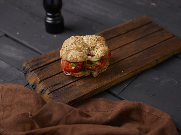 Vegetable sandwich on a seeded croissant on a wooden board with a brown cloth napkin, black background